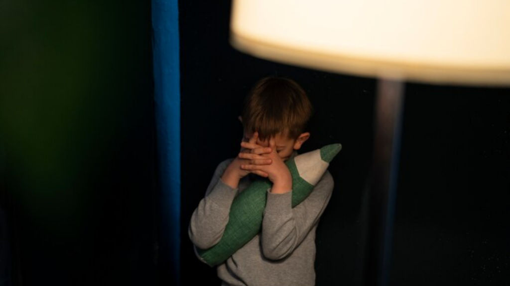 Menino em um quarto escuro cobre os olhos com as mãos enquanto abraça uma almofada verde em formato de lápis, ao lado de um abajur aceso