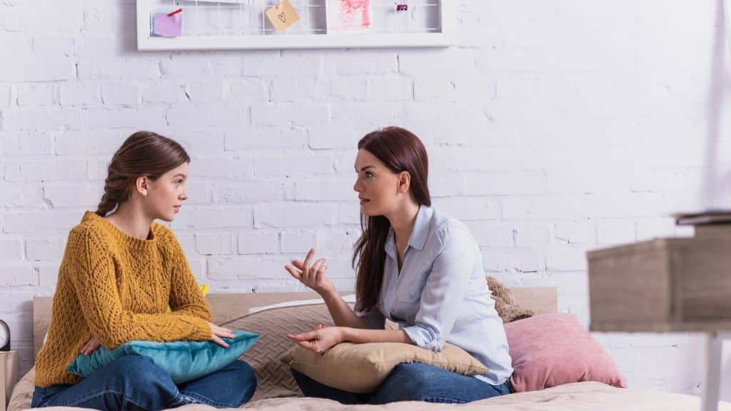 Mãe e filha adolescente sentadas na cama, uma de frente para a outra, segurando almofadas enquanto conversam em um quarto de paredes brancas Filho adolescente não conversa