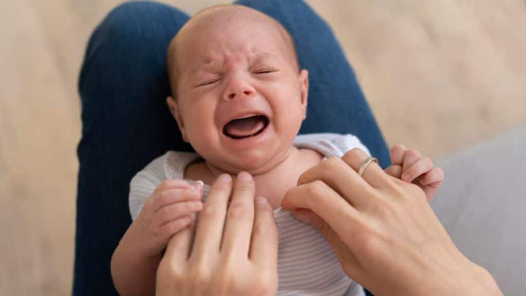 Bebê chora deitado no colo de um adulto, que segura suas mãos