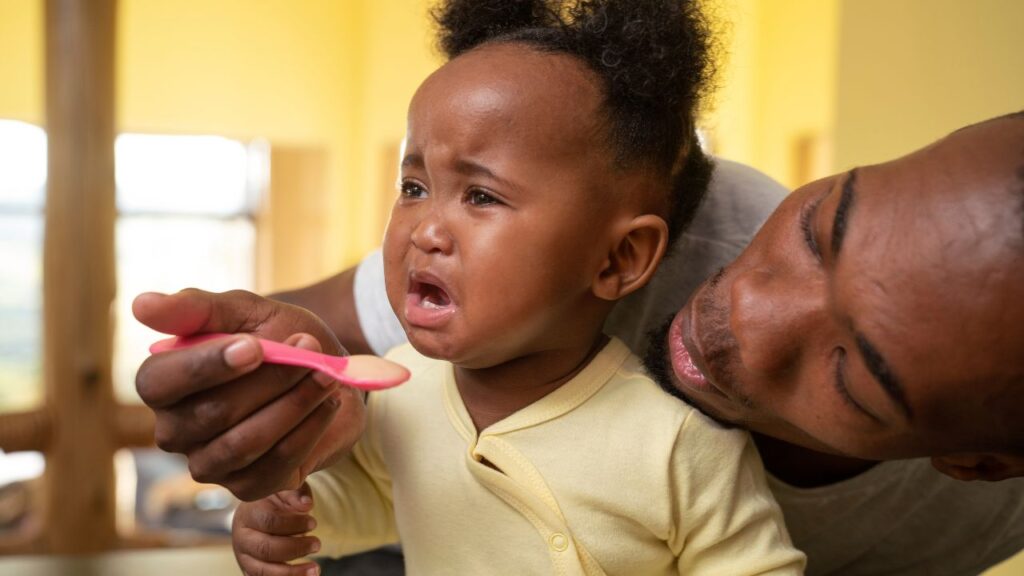 Bebê chora enquanto o pai a segura no colo e oferece comida com uma colher