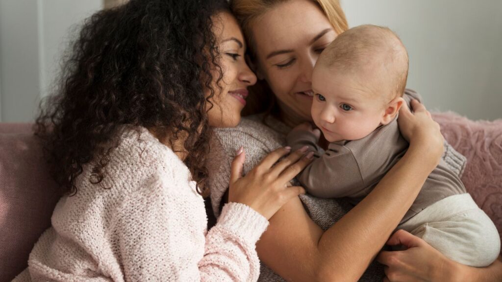 Duas mães sorridentes abraçam um bebê, sentadas em um sofá rosa