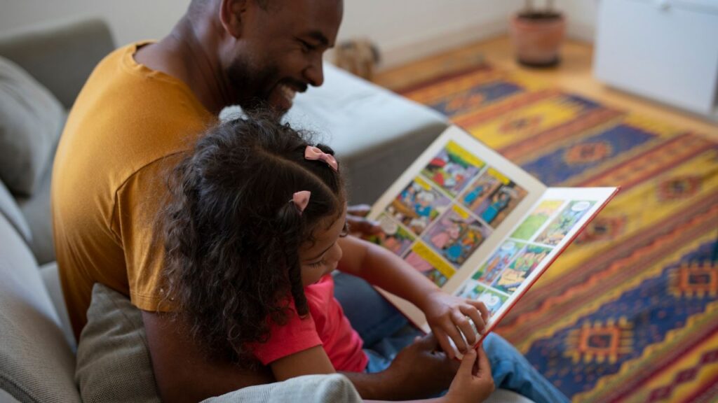 Homem sorridente sentado no sofá com uma menina no colo, os dois folheando um livro em quadrinhos colorido